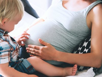 Image Of A Pregnant Mother Holding Her Baby Bump, While A Toddler Is Sitting On Her Thighs, While They Both Admire The Baby Bumps