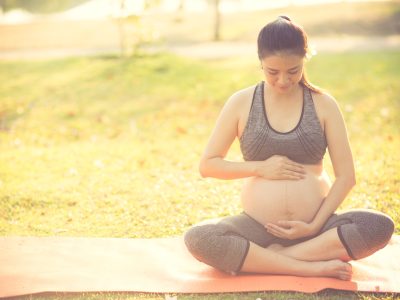Image Of A Pregnant Mother Holding Her Baby Bump With Both Hands, While Sitting On A Yoga Mat In The Park, Woman Is Wearing Yoga Clothing